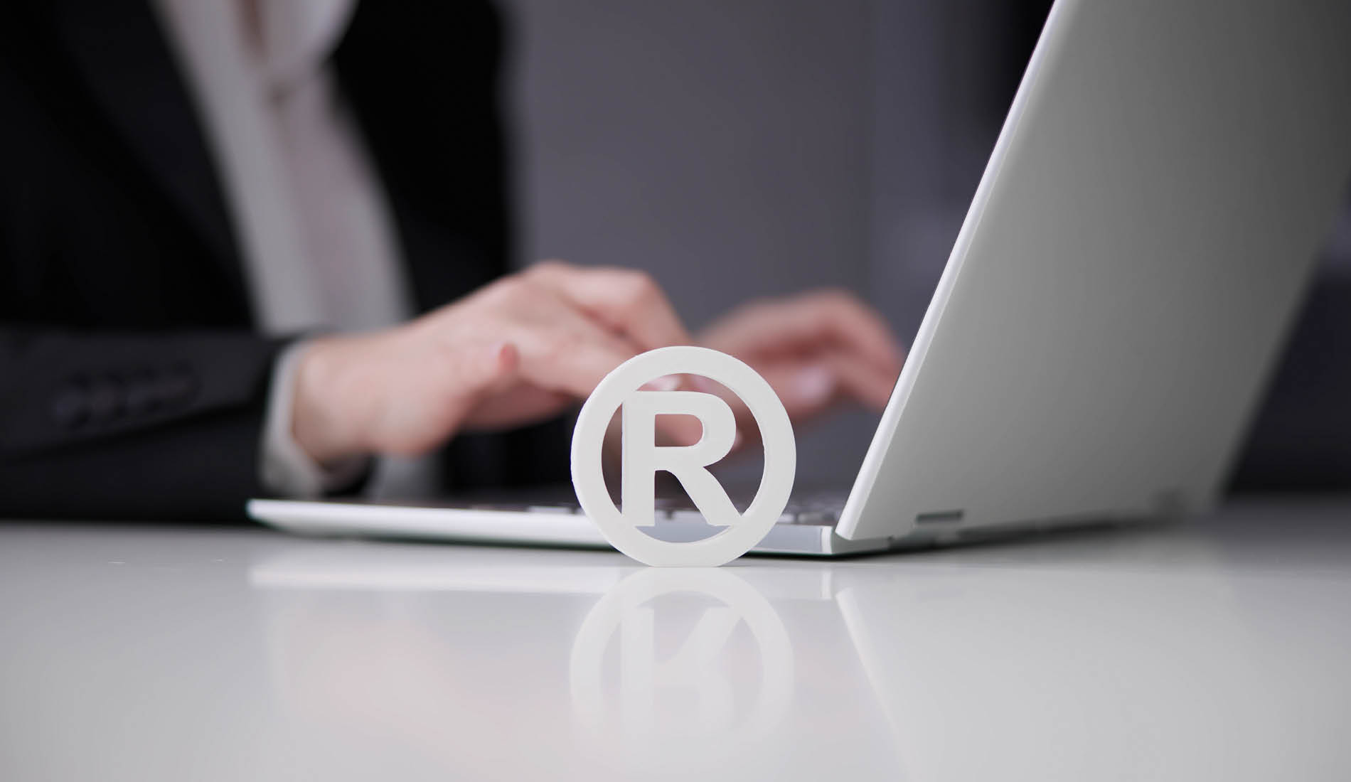 Person arbeitet am Laptop, im Vordergrund steht ein 3D-Registrierungszeichen als Symbol für Markenschutz und Namensfindung.