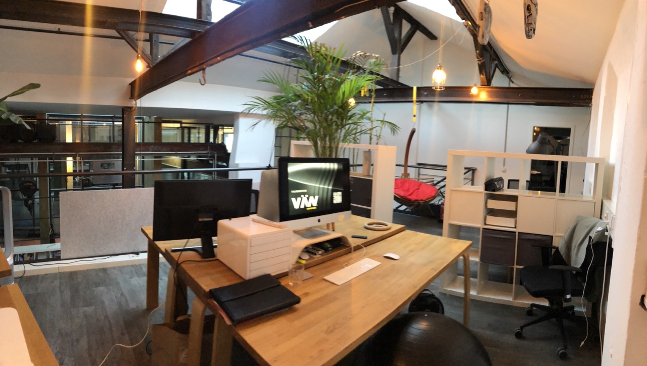 Sendesignal Designstudio – Büro und Arbeitsumgebung Modernes Designbüro mit Holzschreibtischen, Pflanzen, iMac-Arbeitsplatz und industriellen Dachbalken im Loftstil.