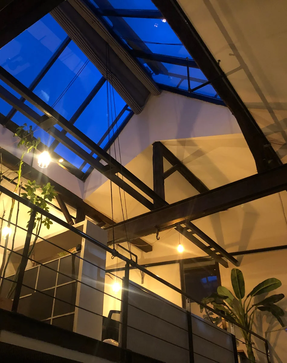 Loft-Büro am Abend mit Glasdach, dunklen Holzbalken, Pendelleuchten und Zimmerpflanzen.